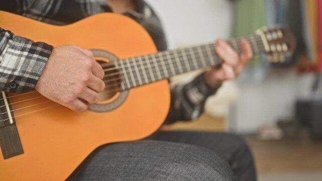 Man playing a classical guitar, fingers on the fretboard and right bare hand strumming chords while seated in a studio; calm concentration.