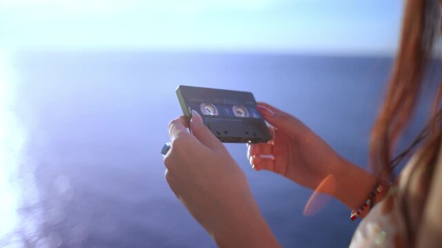 Young woman holds an audio cassette tape in both hands against a bright ocean background, digital detox listening using an analog device