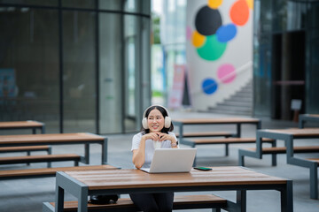 Fototapeta premium A smiling young woman wearing headphones works remotely on her laptop, studying and communicating online while enjoying music—an image that reflects the use of technology, work efficiency, and 