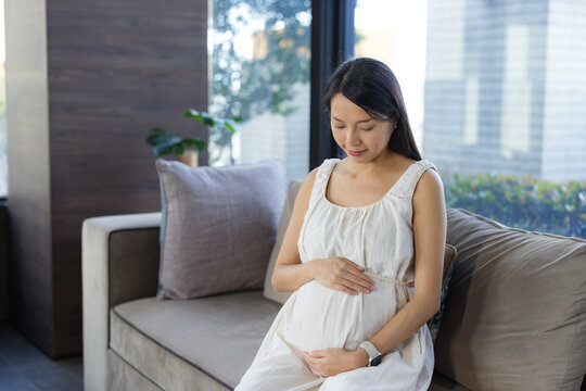 Pregnant woman sitting on a living room sofa gently touching her belly