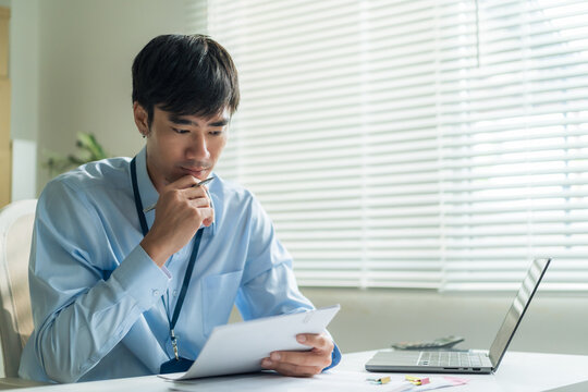 Office worker holding pen and checking report document thoughtfully at desk.