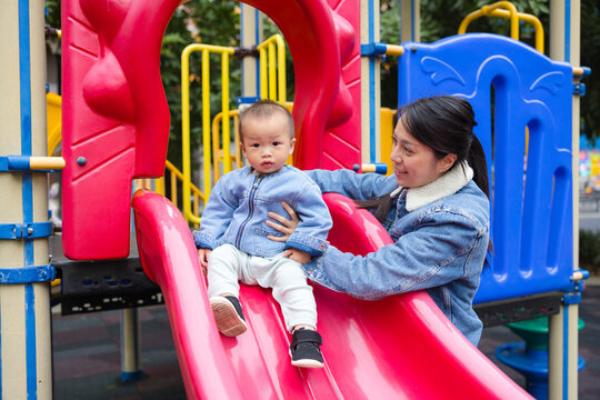 Mother helping her toddler son go down a red slide at an outdoor playground