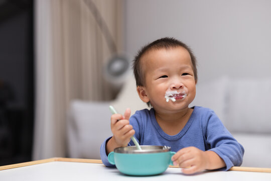 Happy toddler boy with food on his face holding a spoon at a table indoors