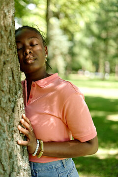 Young adult African American woman leaning against tree trunk with eyes closed in park, practicing ecotherapy. Image suitable for mental health and outdoor wellness campaigns, vertical