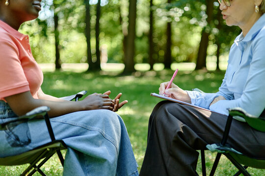 Middle aged Caucasian female therapist taking notes during outdoor counseling session with young adult African American woman in park, promoting mental health services