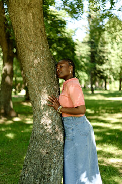 Young adult African American woman leaning against tree trunk with closed eyes in park, practicing ecotherapy, representing mental wellbeing and outdoor psychotherapy, vertical