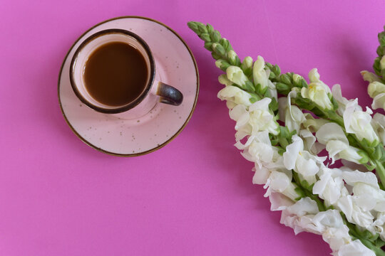 Pink background, cup of coffee, delicate white flowers. Coffee steams gently beside blooming white blossoms. Simple, calm, and inviting composition.