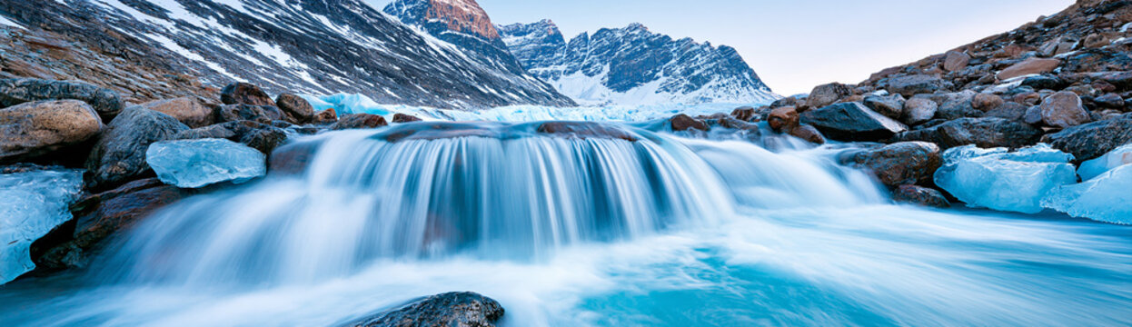 A serene icy waterfall flows rapidly among rocks, with snow-covered mountains in the background under a clear sky.