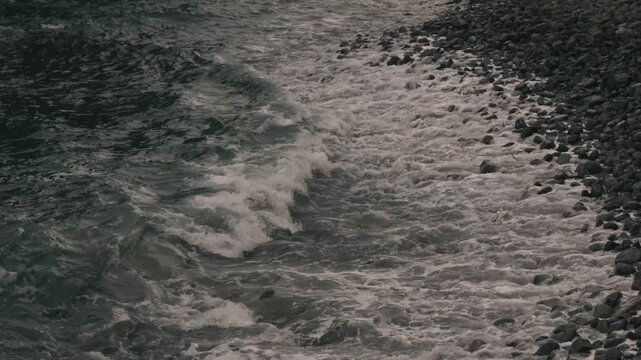 Waves crashing violently against numerous rounded stones along a rocky shore
