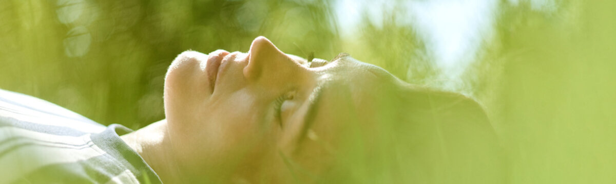 Header of Caucasian young adult woman lying on grass with eyes closed during outdoor psychotherapy session. Image suitable for mental health recovery and wellness campaign, close up