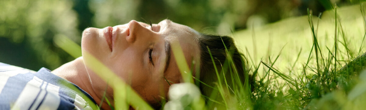 Caucasian woman lying on green grass with eyes closed during outdoor psychotherapy session, representing mental wellbeing and suitable for healthcare marketing, close up header