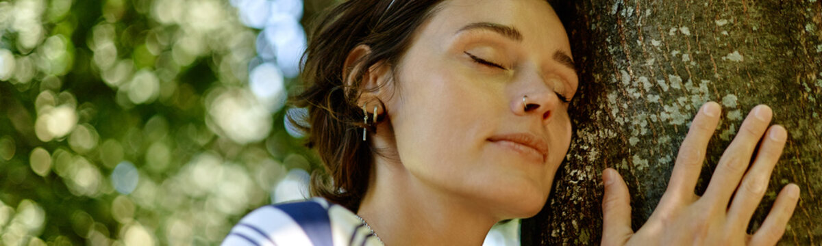 Young adult Caucasian woman leaning head on tree trunk with closed eyes during ecotherapy session in park, representing mental wellbeing and nature therapy, close up header
