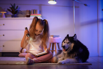 Childhood imagination and bedtime reading, little girl with flashlight reading a book to her husky dog in a cozy bedroom at night © Pixel-Shot