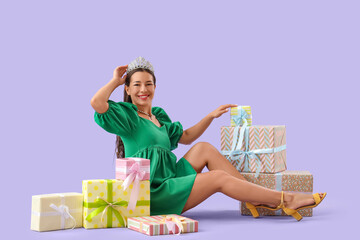 Naklejka premium Birthday celebration and gift giving, happy woman wearing a tiara sitting with wrapped presents on a purple background for holiday shopping concepts