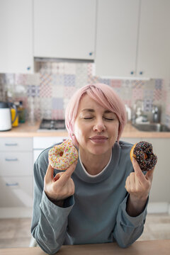 Mature woman holds two colorful donuts with closed eyes, surrendering to intense PMS cravings and sugar addiction after trying to avoid sweet pastries and maintain dietary self-control.