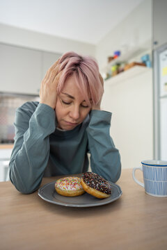 Mature woman sits distressed before colorful donuts, struggling with PMS, sugar addiction and pastry cravings while trying to avoid sweets and maintain self-control during difficult withdrawal.