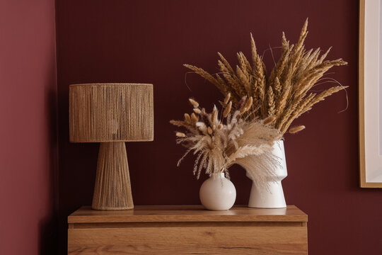 Interior design and home decor, modern table lamp with dried pampas grass in vases on wooden console table against dark red wall in living room