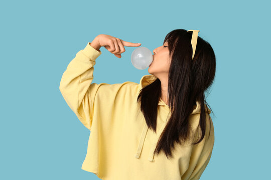 Pretty young Asian woman blowing bubble gum and pointing at it on blue background