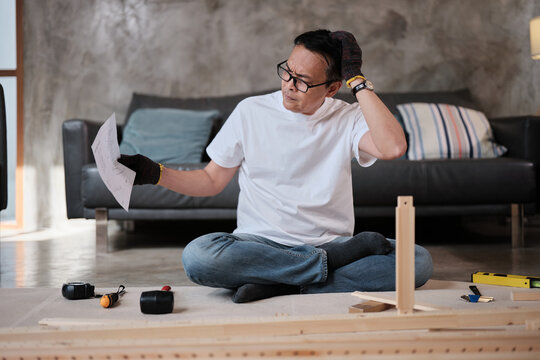 Asian mature man self-assembling flat-pack furniture with DIY tools and instruction guide, confused with wooden parts, home improvement, craft installation, hobby workshop, handyman active lifestyle.