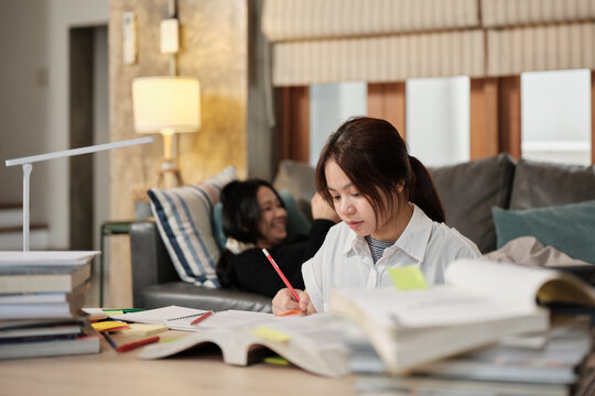Two Asian teenage students learning at home together, a girl concentrates on a pile of books, her friend relaxes on sofa, self-studies, reading textbooks, homeschooling education together lifestyle.