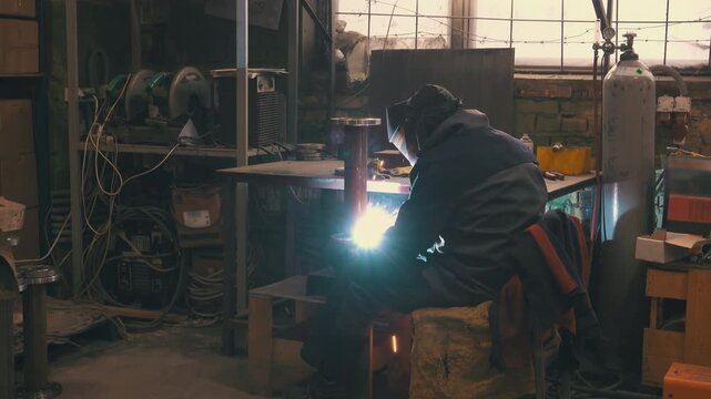 Welder Working in Metal Fabrication Workshop Wide Shot | Bright Welding Flash Illuminating Industrial Garage Interior | Man Welding Steel Pipe Flange at Workbench