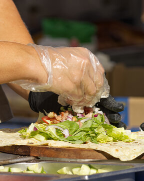 A gloved hand sprinkles crumbled cheese over a fresh flatbread wrap piled with lettuce, arugula, tomato and diced savory toppings at a Naplavka market stall in Prague.

