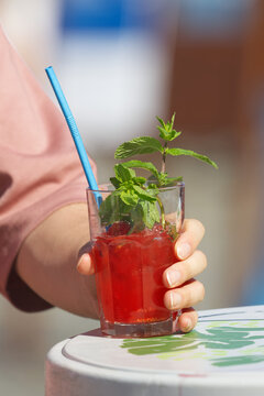 A hand holds a vibrant iced red fruit drink garnished with fresh mint sprigs, ice cubes and a blue straw on a hand-painted cafe table at the sunny Naplavka market in Prague.
