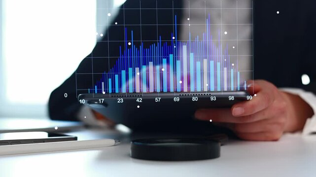 A business person is sitting at a desk while looking at data on a tablet. Colored graphs and charts show information on the screen. The setting is likely an office during daytime.
