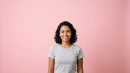 Fototapeta premium Young woman with dark curly hair and a friendly smile wearing a grey t-shirt against a pink background