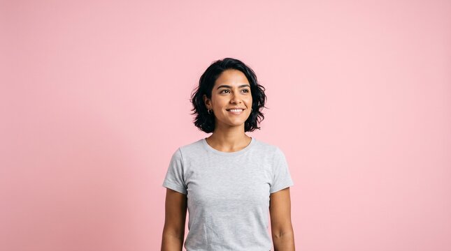 Young woman with dark wavy hair and a grey t-shirt smiling and looking to the side against a pink background