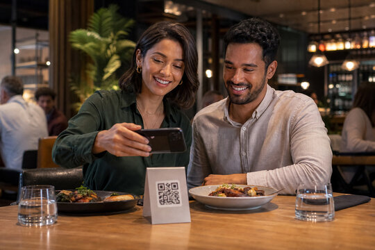 Diverse Young Couple Scanning a Tabletop QR Code with a Smartphone to Complete a Cashless Payment After Dinner in a Stylish Evening Bistro
