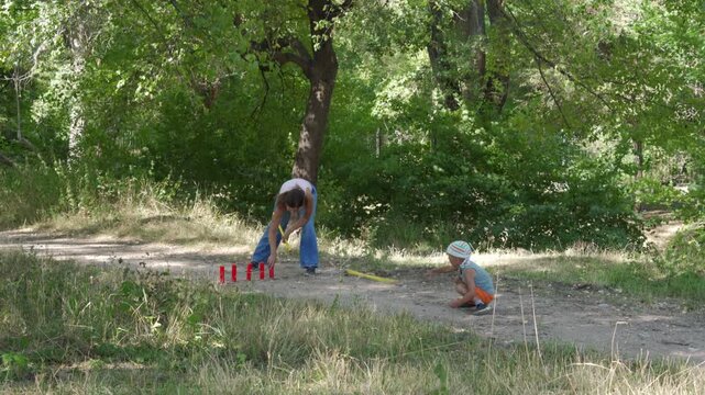 A loving mother and her happy son joyfully play a game of gorodki on a dirt path. They share a cheerful, bonding moment. A peaceful green forest surrounds them.