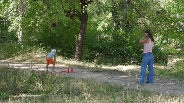 A loving mother and her happy son joyfully play a game of gorodki on a dirt path. They share a cheerful, bonding moment. A peaceful green forest surrounds them.