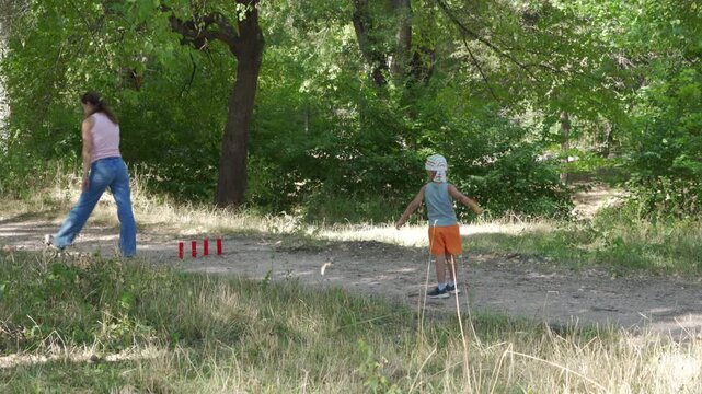 A loving mother and her happy son joyfully play a game of gorodki on a dirt path. They share a cheerful, bonding moment. A peaceful green forest surrounds them.