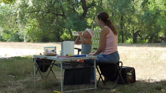 A caring mother watches her thirsty son drink from a bottle at a picnic table. They share a peaceful, content moment together. A sunny green park surrounds them.