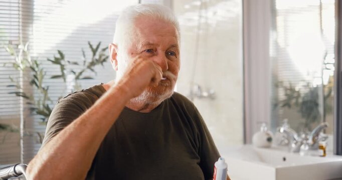 Senior man in a wheelchair applies toothpaste to a toothbrush and brushes his teeth during his morning bathroom routine at home.