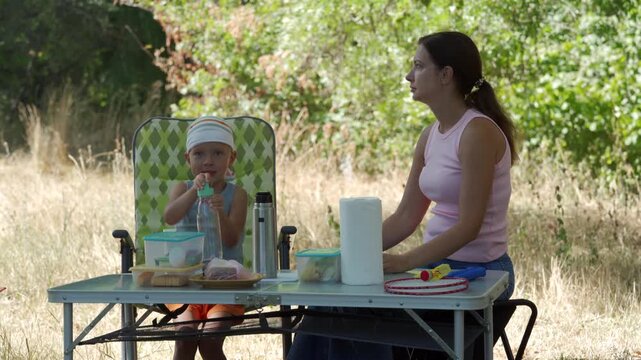 Young child sits at folding table eating snack while mother stands nearby arranging paper towel and juice during casual picnic setup in grassy park, surrounded by trees, sunshine, calm family