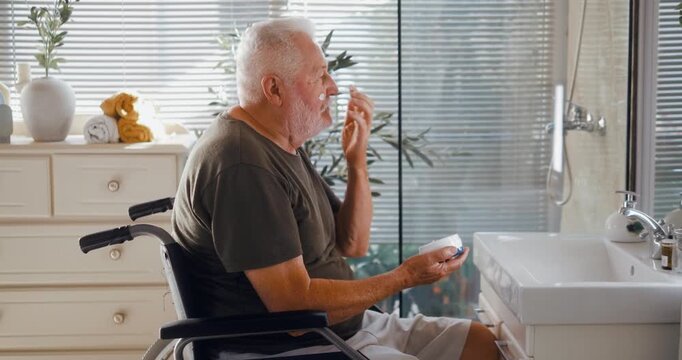 Side view of a senior man in a wheelchair applying face cream while looking in the bathroom mirror during his morning skincare routine at home.