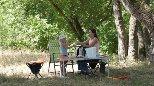 A helpful mother and her happy son organize picnic containers at a folding table. They share a loving, cheerful moment while packing. A peaceful green forest surrounds them.