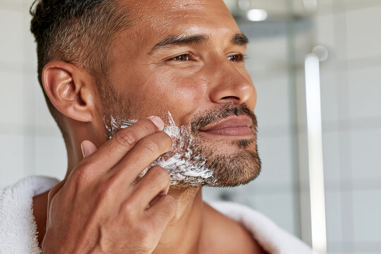 Man shaves face with white foam. Focused, calm, and serene expression. Bright bathroom with modern tiles. Ideal for men's grooming, wellness, or daily routine content