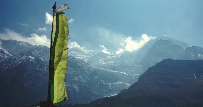 Annapurna Circuit, Manang, Nepal, close-up view of a yellow Buddhist prayer flag waving on a stone pillar with snow-capped mountain peaks in the background