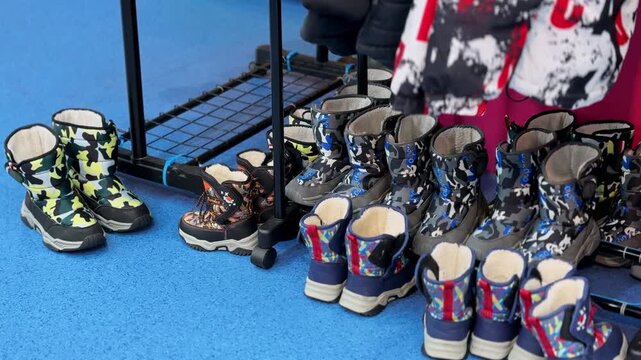 A row of warm children's winter snow boots with camouflage patterns lined up on a blue floor indoors. The footwear sits under a clothing rack in a winter amusement rental shop under bright lighting.