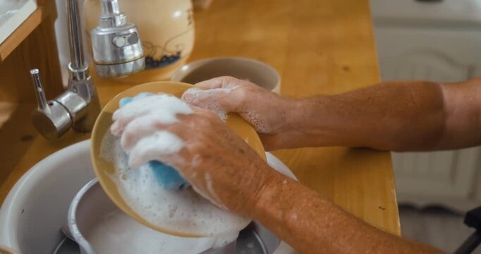 Close-up of a senior man's hands washing a plate with a sponge and soap foam in the kitchen sink during a daily routine at home.