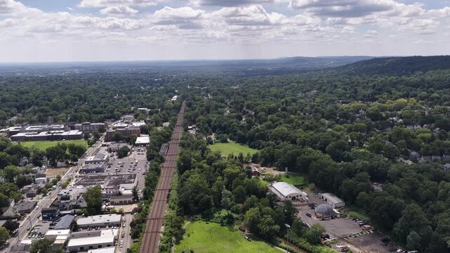 The drone looks along the railway corridor, then tilts downward and advances toward the tracks, open green space and wooded land near Maplewood and South Orange.