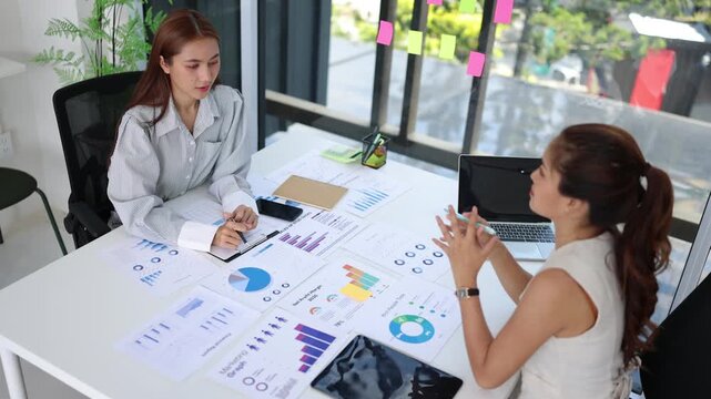 Two Asian businesswomen discussing financial reports and analyzing market data in a modern office meeting.