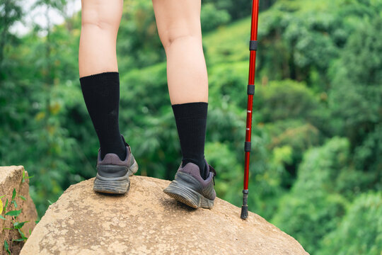 Legs of a person standing on mountain rock wearing black shoes and socks holding red trekking pole during hike