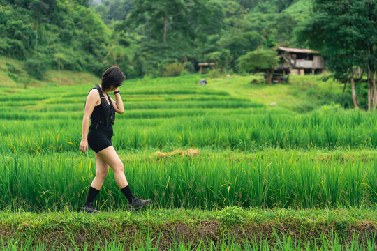 Young woman walking along lush green rice field pathway with traditional rural landscape and wooden house in background