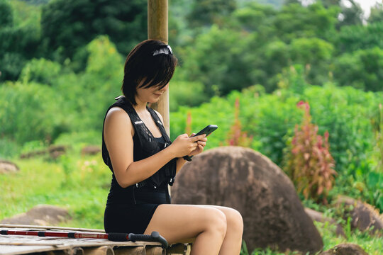 Young woman in black outfit sitting outdoors on wooden bench looking at smartphone beneath rustic shelter in nature