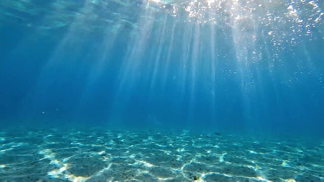 Stunning underwater view of clear blue ocean with bright sun rays piercing through the surface, illuminating the sandy seabed.