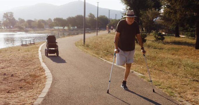 Senior amputee walks independently with crutches beside his electric wheelchair on a lakeside path, showing active aging, mobility and everyday physical activity.